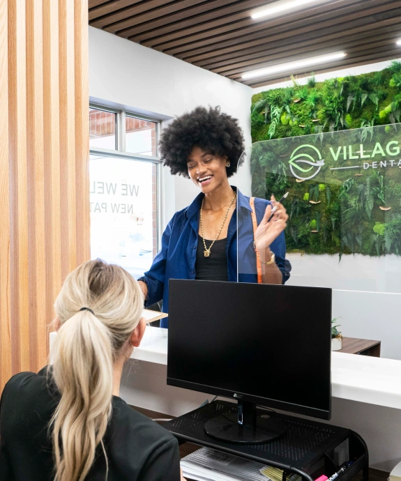 Dental receptionist greeting patient at front desk in modern office with green plant wall.