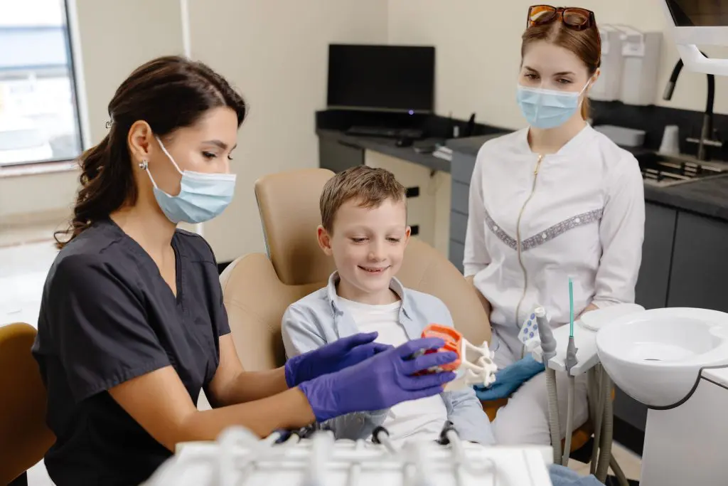 A young child reclines in a dental chair, while a mother sits beside them offering comfort; a gloved dentist leans in to examine the child’s teeth.
