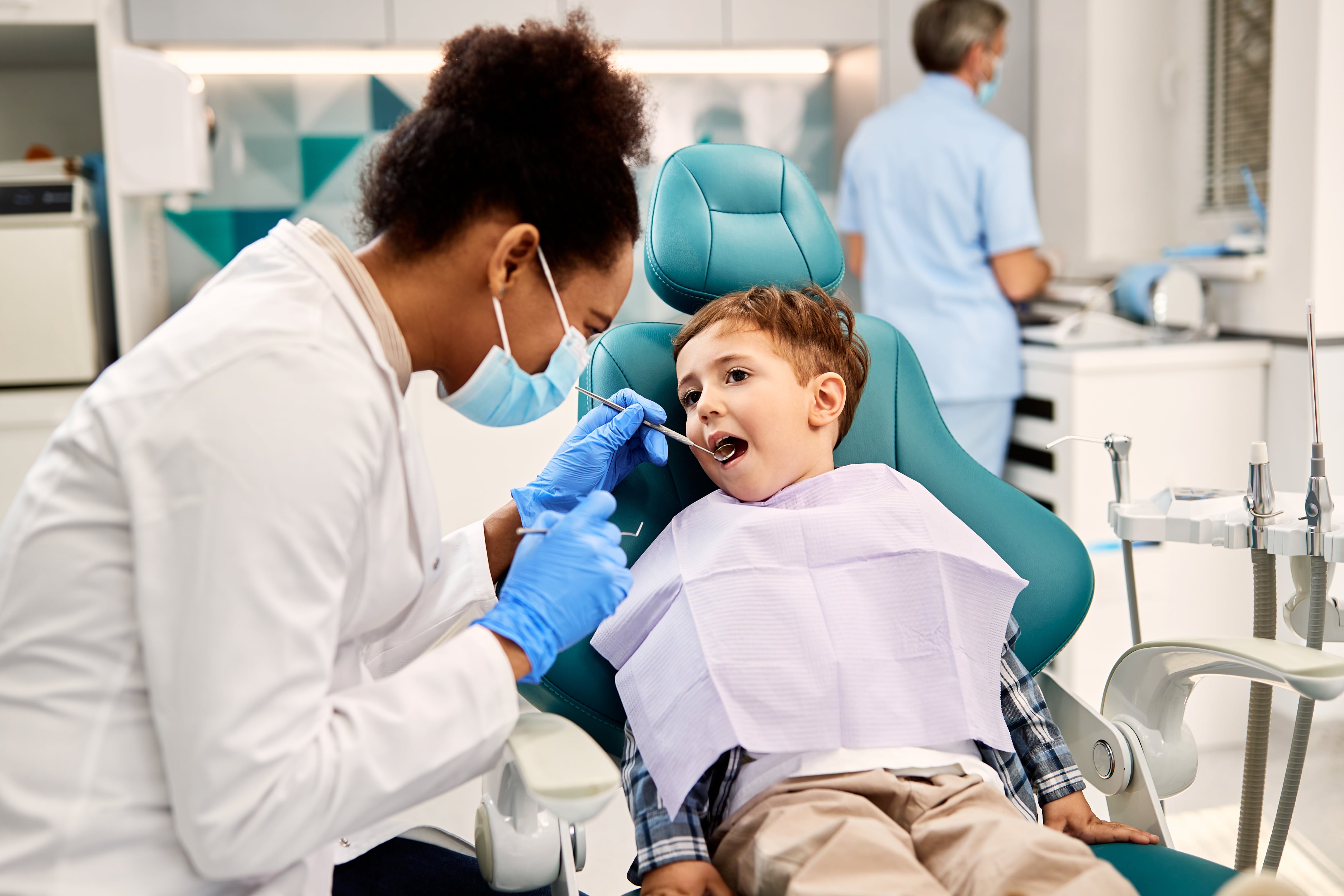 A pediatric dentist examining a smiling child’s teeth in a brightly lit dental clinic.