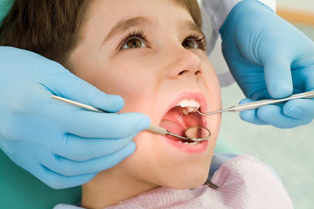A smiling toddler reclines in a dental chair with mouth open, while a pediatric dentist wearing gloves gently examines their teeth.