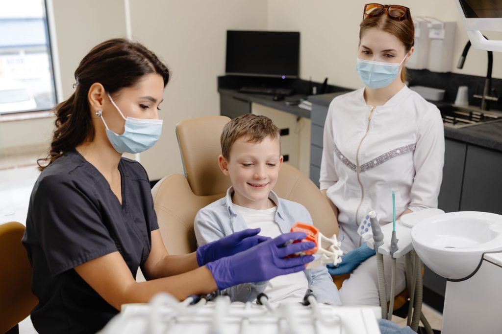 A young child reclines in a dental chair, while a mother sits beside them offering comfort; a gloved dentist leans in to examine the child’s teeth.